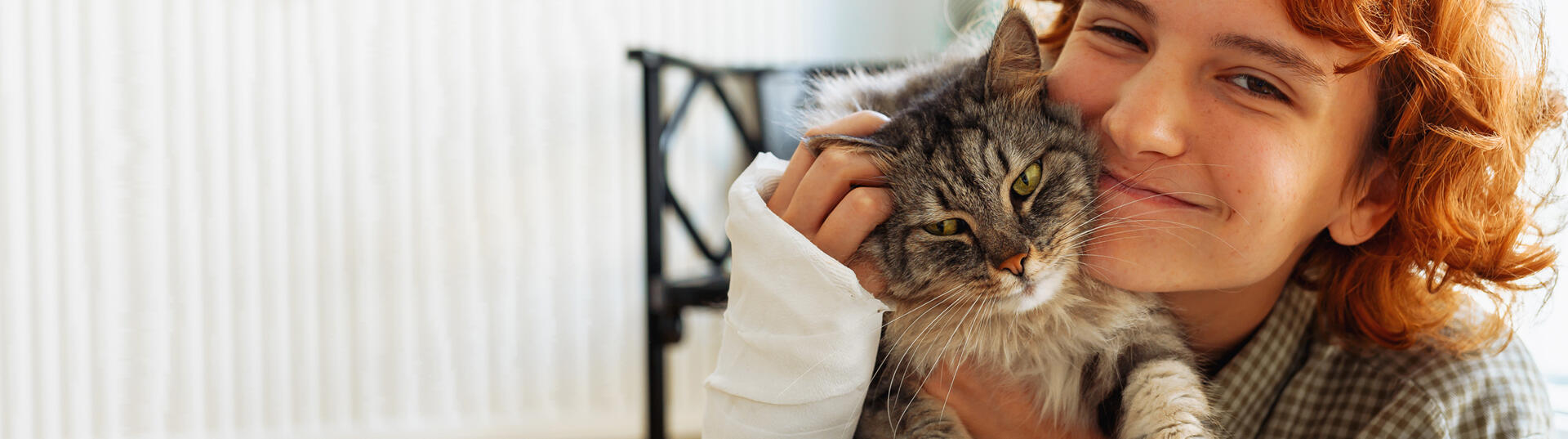 teen girl with curly red hair, broken arm in cast, sits on floor of house, hugging favorite fluffy cat.